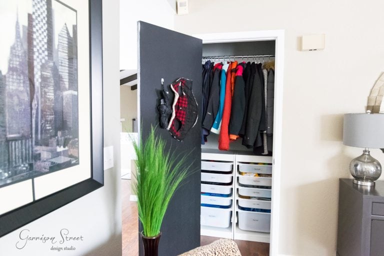 A closet with an open door reveals coats of various colors hanging on a rod. Below are white IKEA drawers, while the door has hats hanging on it. In the foreground, a framed cityscape image and a green plant highlight the organization of this stylish space.