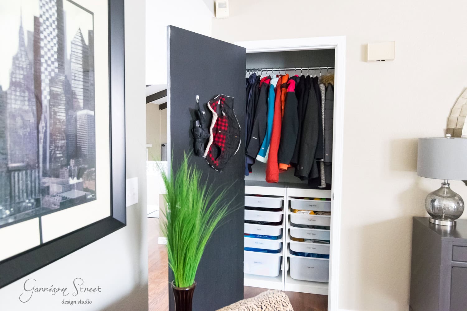 A closet with an open door reveals coats of various colors hanging on a rod. Below are white IKEA drawers, while the door has hats hanging on it. In the foreground, a framed cityscape image and a green plant highlight the organization of this stylish space.