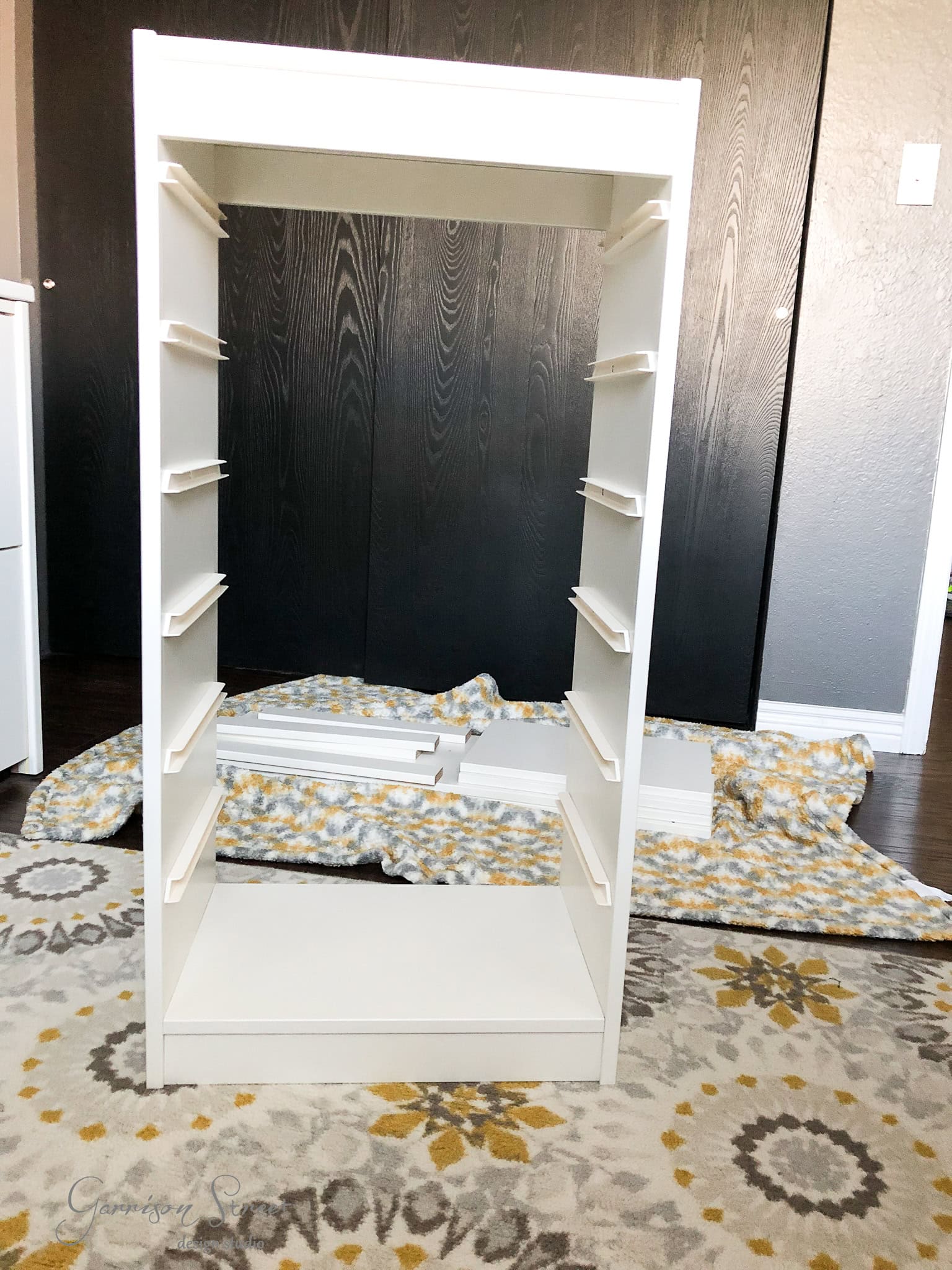A partially assembled white IKEA bookshelf stands on a patterned rug, with other assembly pieces scattered on the floor. Ideal for organization, it leans against a dark wall, waiting to transform any space.