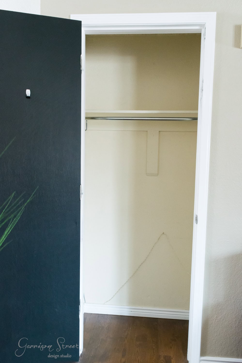 An open Ikea closet with a black door reveals an empty interior, featuring a single shelf and hanging rod. A visible crack in the lower corner of the wall adds character, while a small plant is partially visible on the left side.