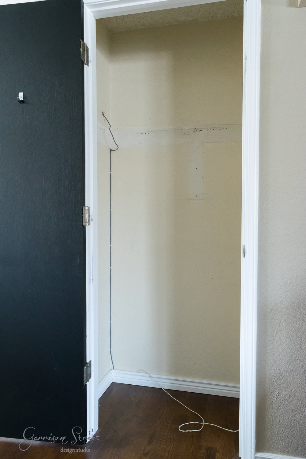 The image showcases an empty beige closet with a black door opened to the left, highlighting organizational possibilities. A white wire dangles against the back wall. The dark wooden floor adds contrast, while part of the right wall is visible, enhancing the Trofast system's potential placement.