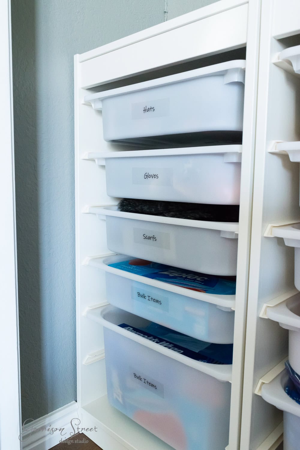A white shelving unit, reminiscent of IKEA's TROFAST series, features labeled plastic bins neatly organized in columns. Labels read "Hats," "Gloves," "Scarfs," and "Bulk Items." The semi-transparent bins reveal folded items inside, set against a grey wall for effortless organization.