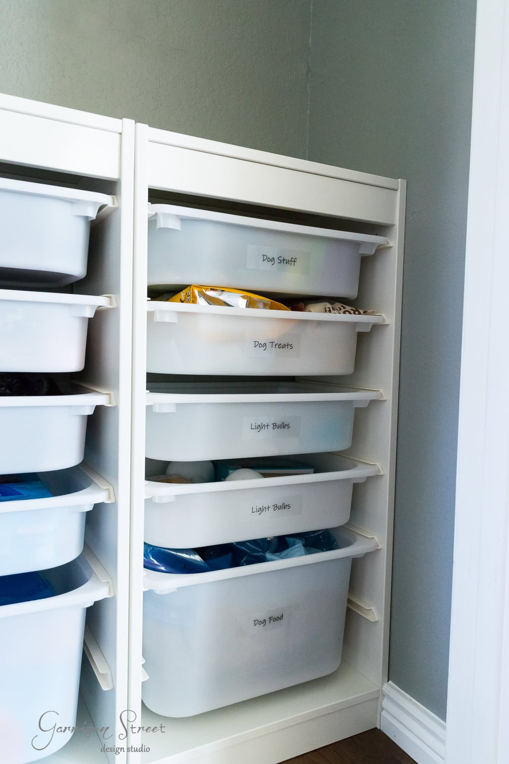 A white Trofast storage unit in a corner with multiple labeled plastic bins offers efficient organization. Labels read "Dog Stuff," "Dog Treats," "Light Bulbs," and "Dog Food." Some bins are slightly open, revealing contents like bags and boxes.
