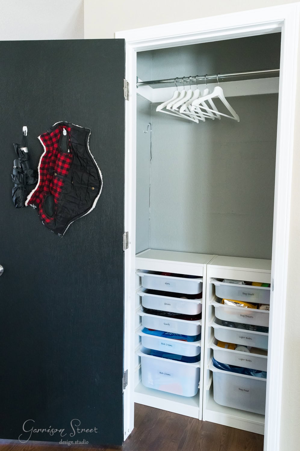 An efficiently organized closet features two columns of labeled IKEA plastic drawers. Above them, white hangers line a rod, while the door displays a plaid garment bag. The wooden floor complements this tidy space.