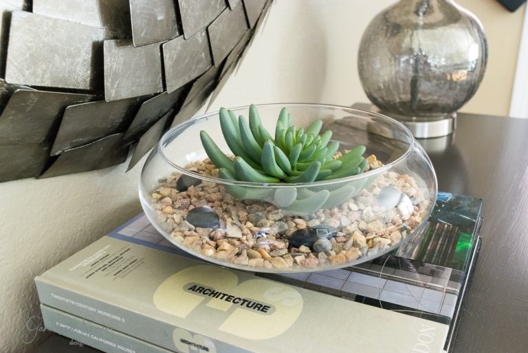 A small glass terrarium with succulent plants rests atop two stacked architecture books. The bowl contains pebbles and small black stones. In the background, a metallic textured decoration and a textured glass lamp complete this elegant DIY setup.