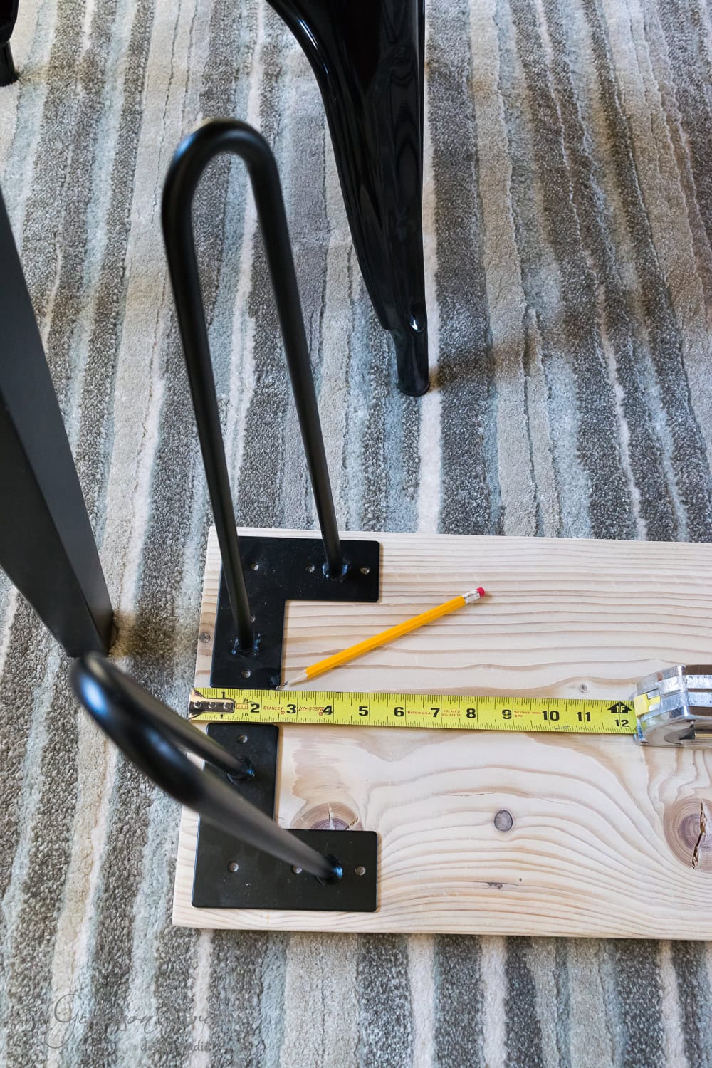 A DIY wooden bench sits on a patterned rug, featuring two sleek black metal hairpin legs. A yellow measuring tape and pencil rest on the plank, ready for precision and creativity.