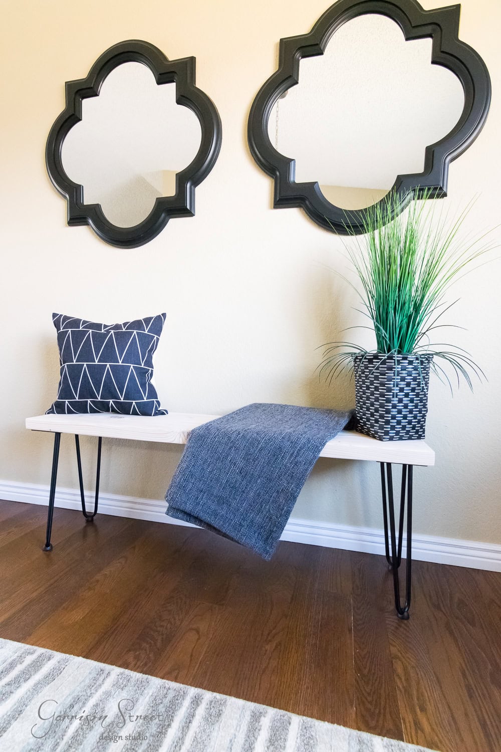 A stylish room showcases a DIY bench with hairpin legs, adorned with a geometric-patterned pillow and a blue throw blanket. A potted plant adds life beside them. Two ornate, quatrefoil mirrors hang on the cream wall above, enhancing the warm wooden floor's appeal.