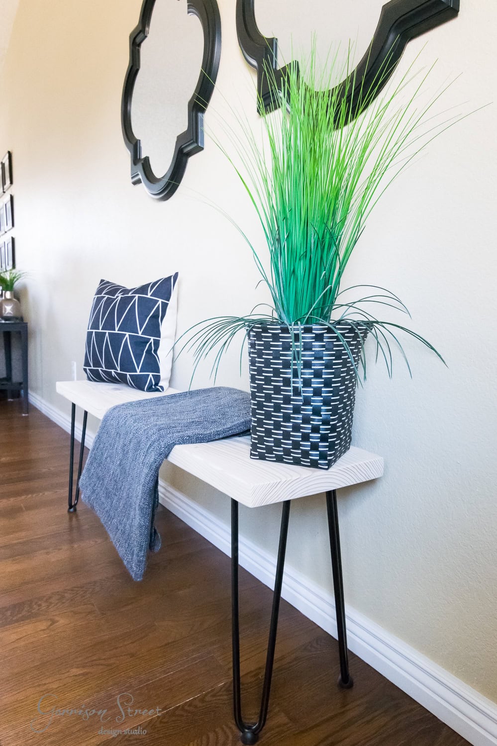 A modern hallway features a wooden bench with sleek hairpin legs. On the bench sits a geometric-patterned pillow, a folded blue blanket, and a potted green plant. Two decorative mirrors hang above on the light-colored wall. Wooden floorboards run below.