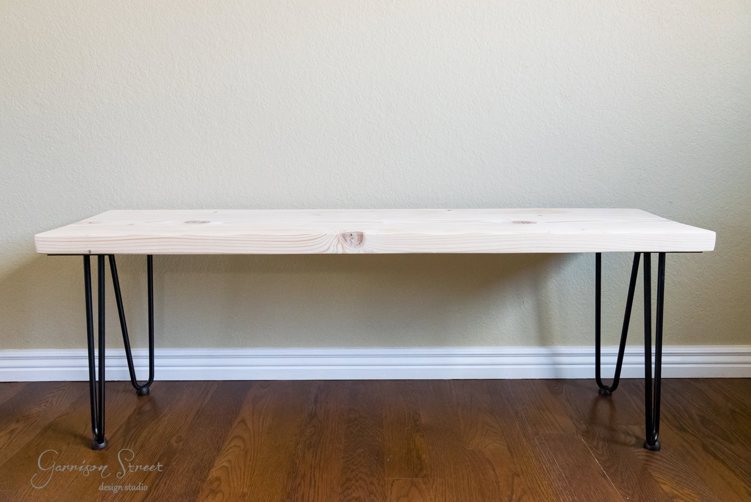 A handmade wooden bench with a light natural finish and sleek black hairpin legs sits elegantly on a hardwood floor against a plain beige wall.