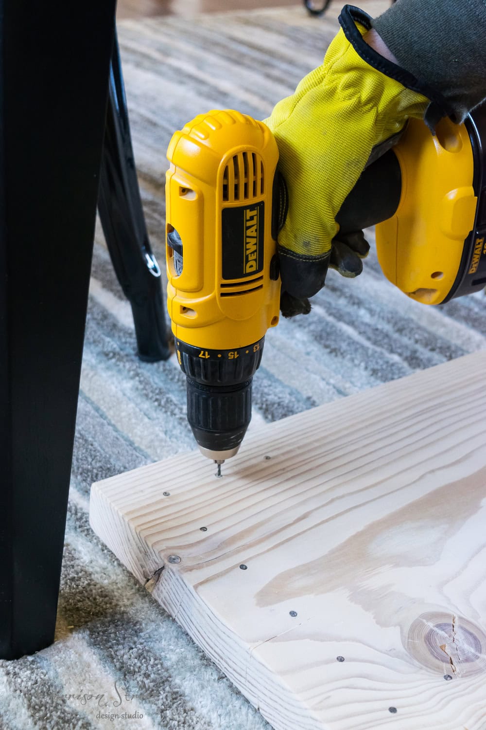 A person wearing yellow gloves is using a yellow cordless drill for a DIY project, screwing into a light wooden plank on a carpeted surface. A black hairpin chair leg is visible to the left.
