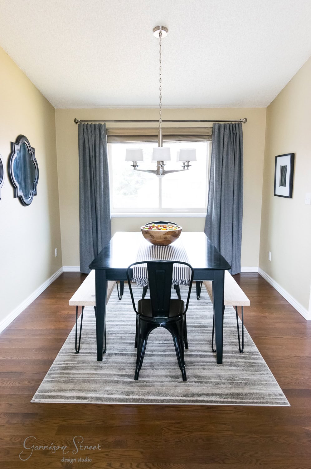 A dining room features a rectangular black table with hairpin legs, flanked by a DIY bench and two black chairs. A fruit bowl sits on the table, while gray curtains hang by the window. A chandelier glimmers above, and wall decorations paired with a striped rug complete the space.