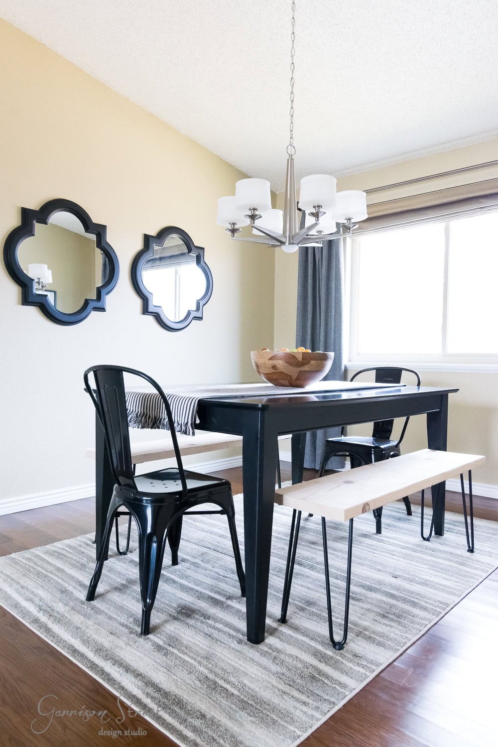 A minimalist dining room features a black table with sleek hairpin legs, complemented by black metal chairs and DIY wooden benches on a striped rug. Two decorative mirrors grace the beige wall alongside a window. A modern chandelier hangs above the table, where a fruit bowl sits gracefully centered.