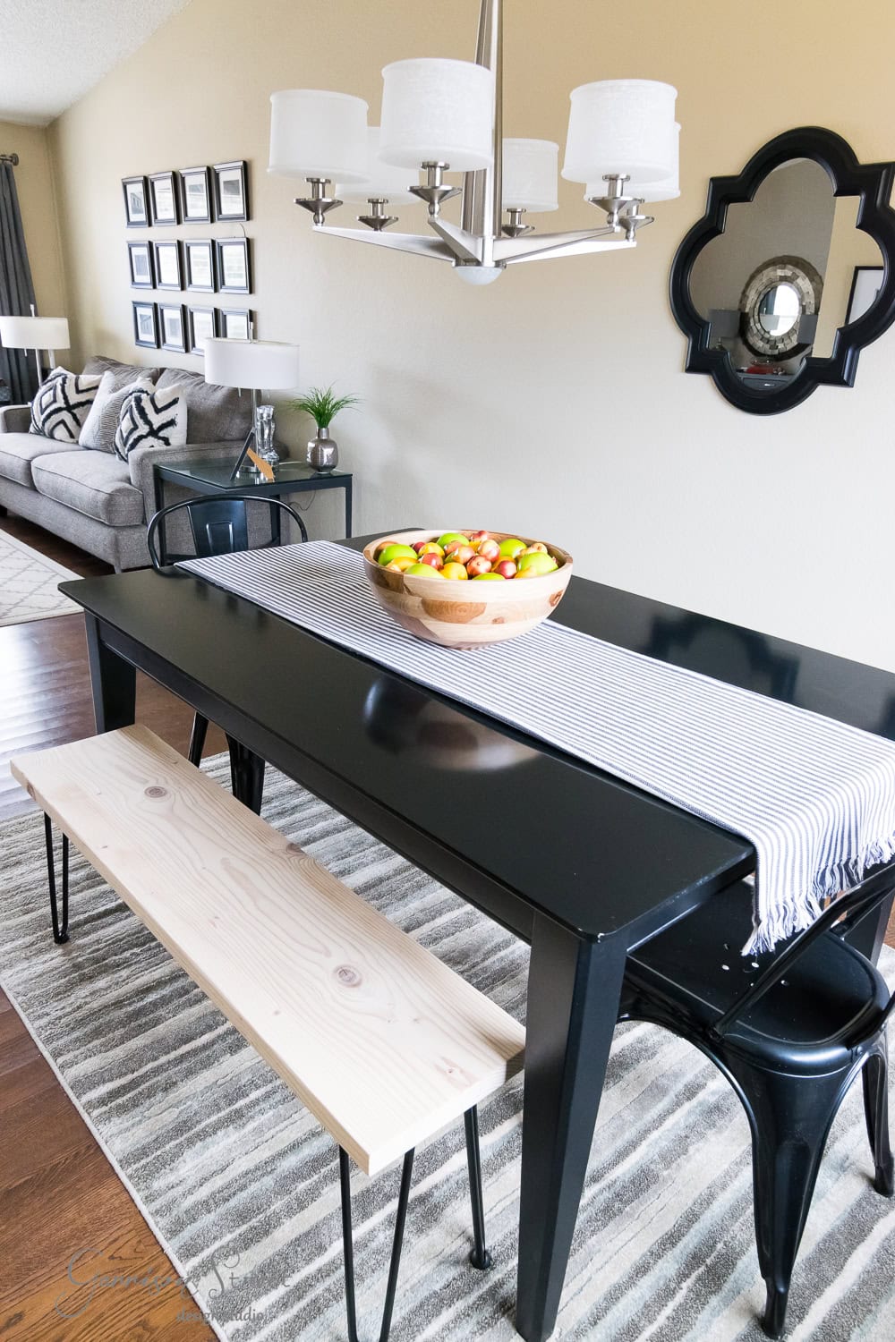 A modern dining area features a black table with sleek hairpin legs, complemented by a light wooden bench and metal chairs. A striped runner and bowl of apples adorn the table. A stylish chandelier hangs above, while a gray sofa, framed photos, and a decorative mirror enhance the neutral walls.