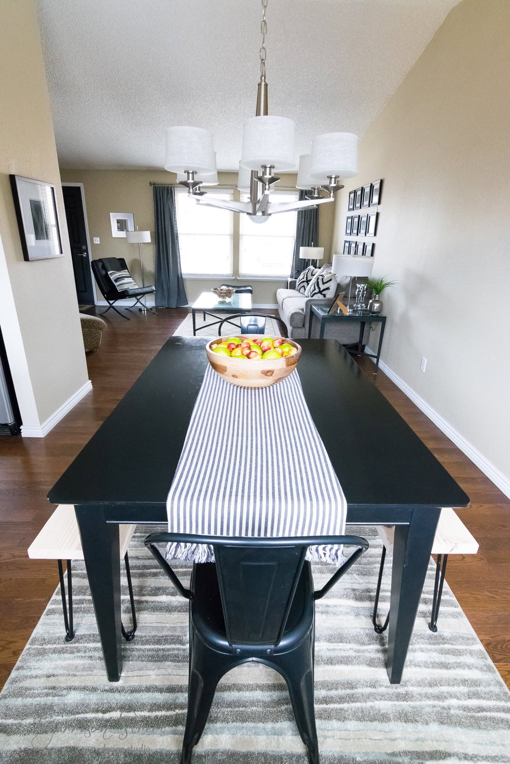 A modern dining room with a black table featuring a striped runner and a bowl of apples. Overhead, a chandelier hangs. A stylish bench with hairpin legs complements the seating in the background living area, which includes framed pictures and large windows. The space boasts wooden flooring and neutral walls.
