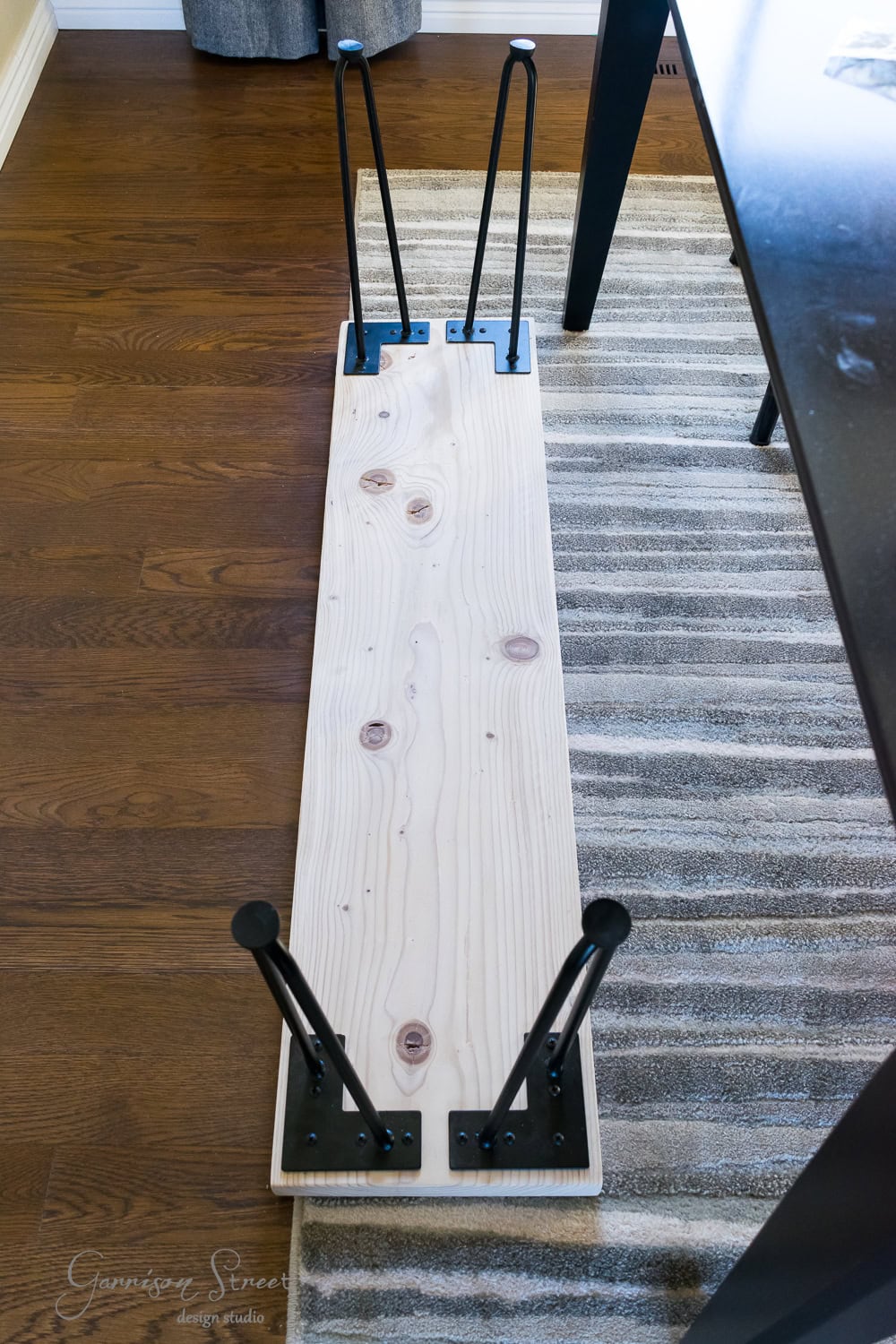 A wooden plank with four black hairpin legs lies upside down on a hardwood floor, partially resting on a gray and white striped rug. This DIY project seems destined to become a stylish bench or table as it's being assembled.