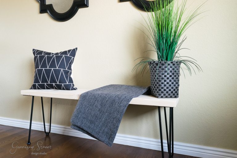 A modern wooden bench with sleek hairpin legs sits against a beige wall. It features a black and white geometric pillow, a textured gray throw blanket, and a tall plant in a black woven pot. Mirrors hang above the bench, adding to the chic DIY aesthetic.