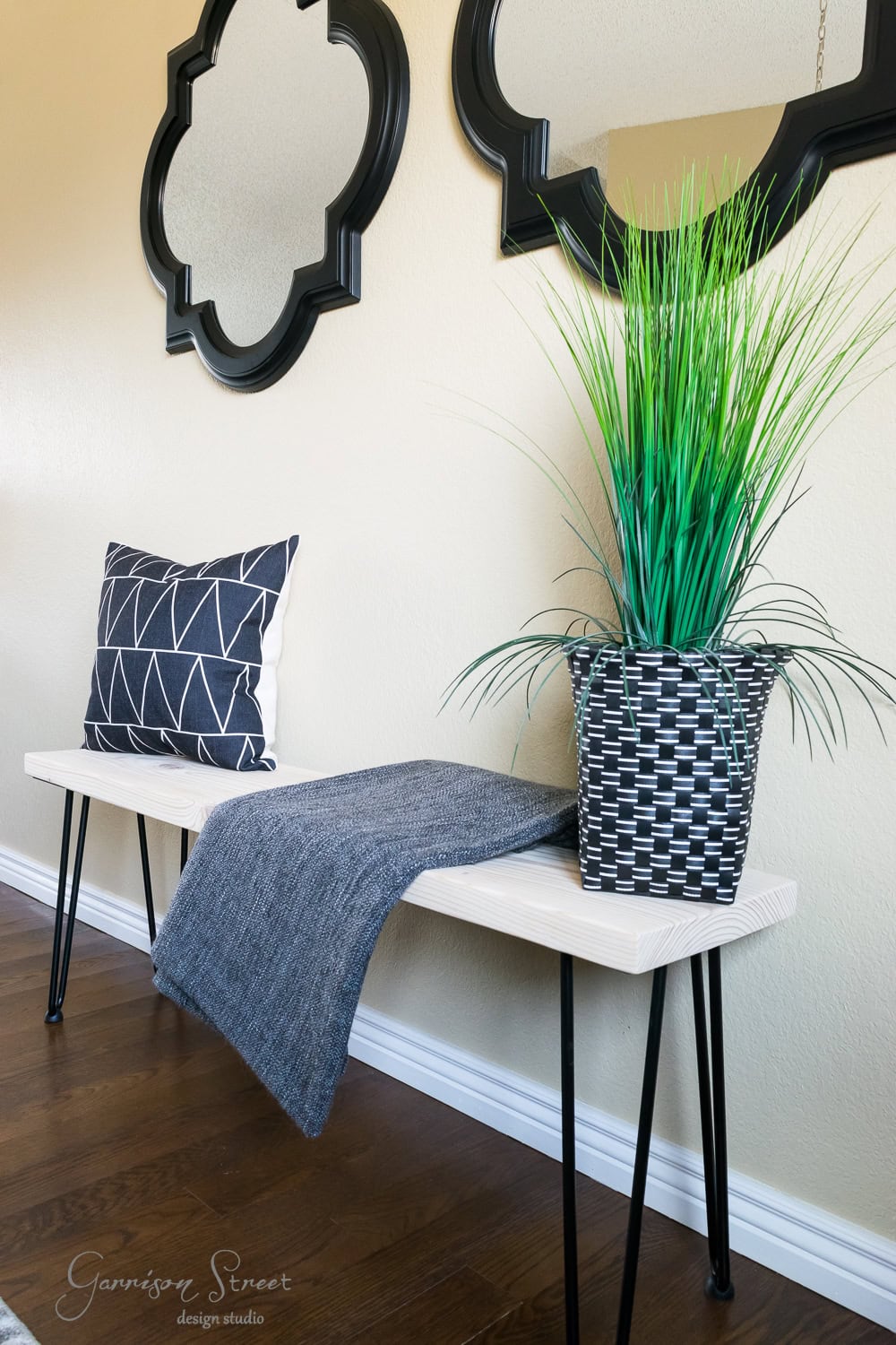 A wooden bench with sleek hairpin legs features a geometric-patterned cushion and a gray blanket. A woven planter holds green grass-like plants. Two decorative mirrors with black frames are mounted on the wall above the bench, complementing the dark wood flooring.