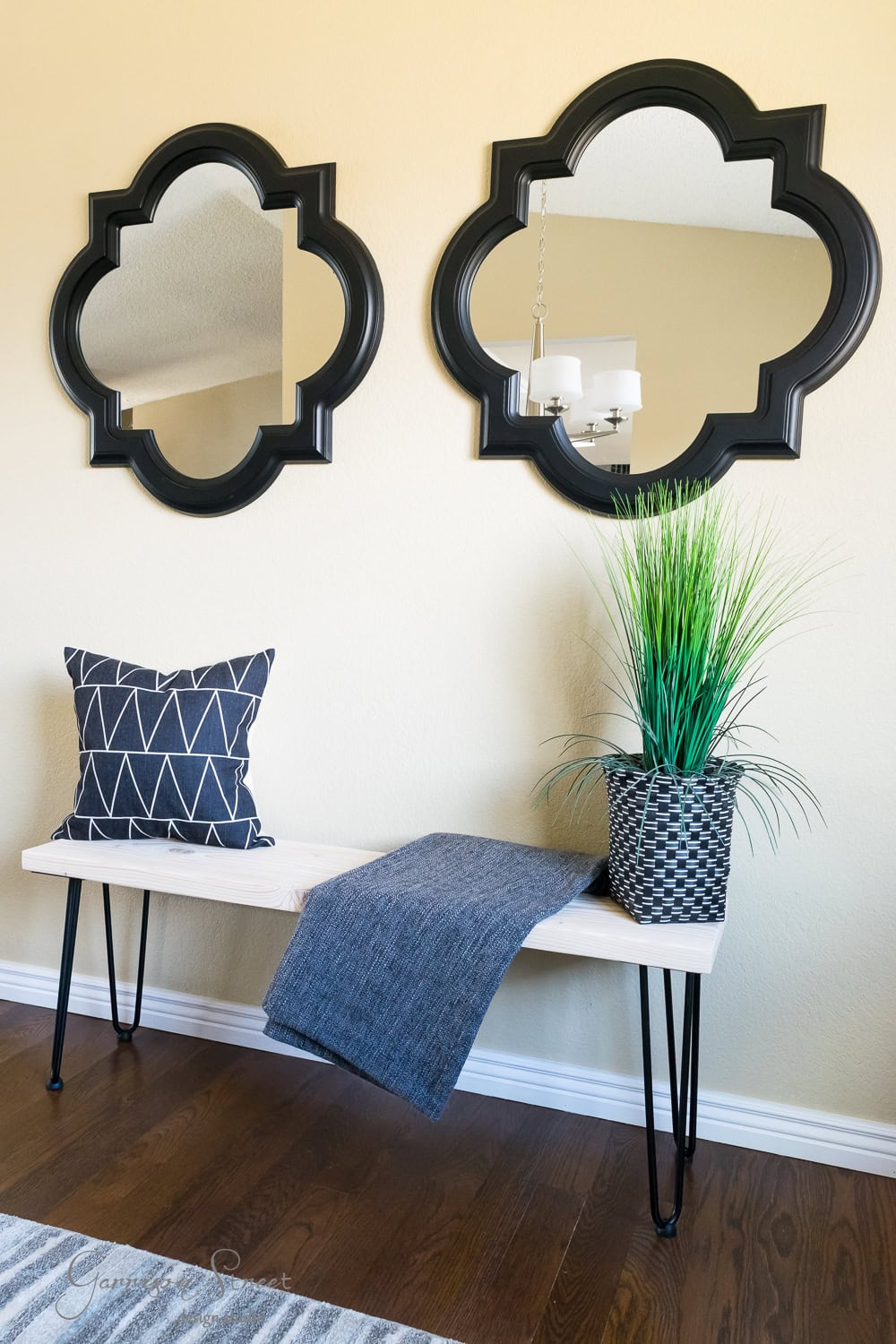 A cozy entryway features a DIY wooden bench with hairpin legs adorned by a geometric-print pillow and a textured blanket. A potted plant sits beside them, while two quatrefoil-shaped mirrors with dark frames grace the beige wall above, reflecting a chandelier.