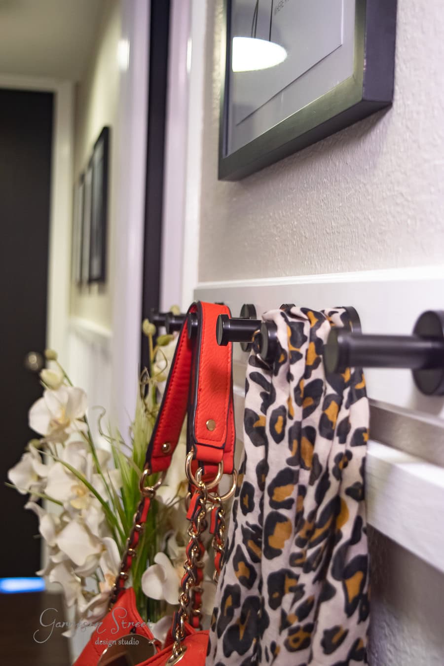 A hallway with a board and batten wall features a black rod holding a red handbag and a leopard print scarf. A bouquet of white flowers is placed nearby, complementing the DIY charm. Framed pictures hang above the rod.