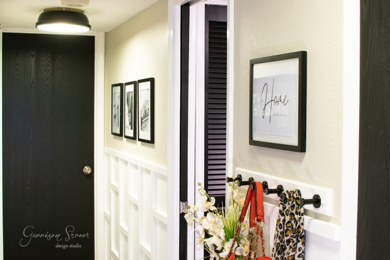 A hallway with black and white decor featuring framed photos on the wall. A "home" sign hangs next to a coat rack adorned with scarves and a bouquet of white flowers in a vase. A black door is visible at the end of the hallway.