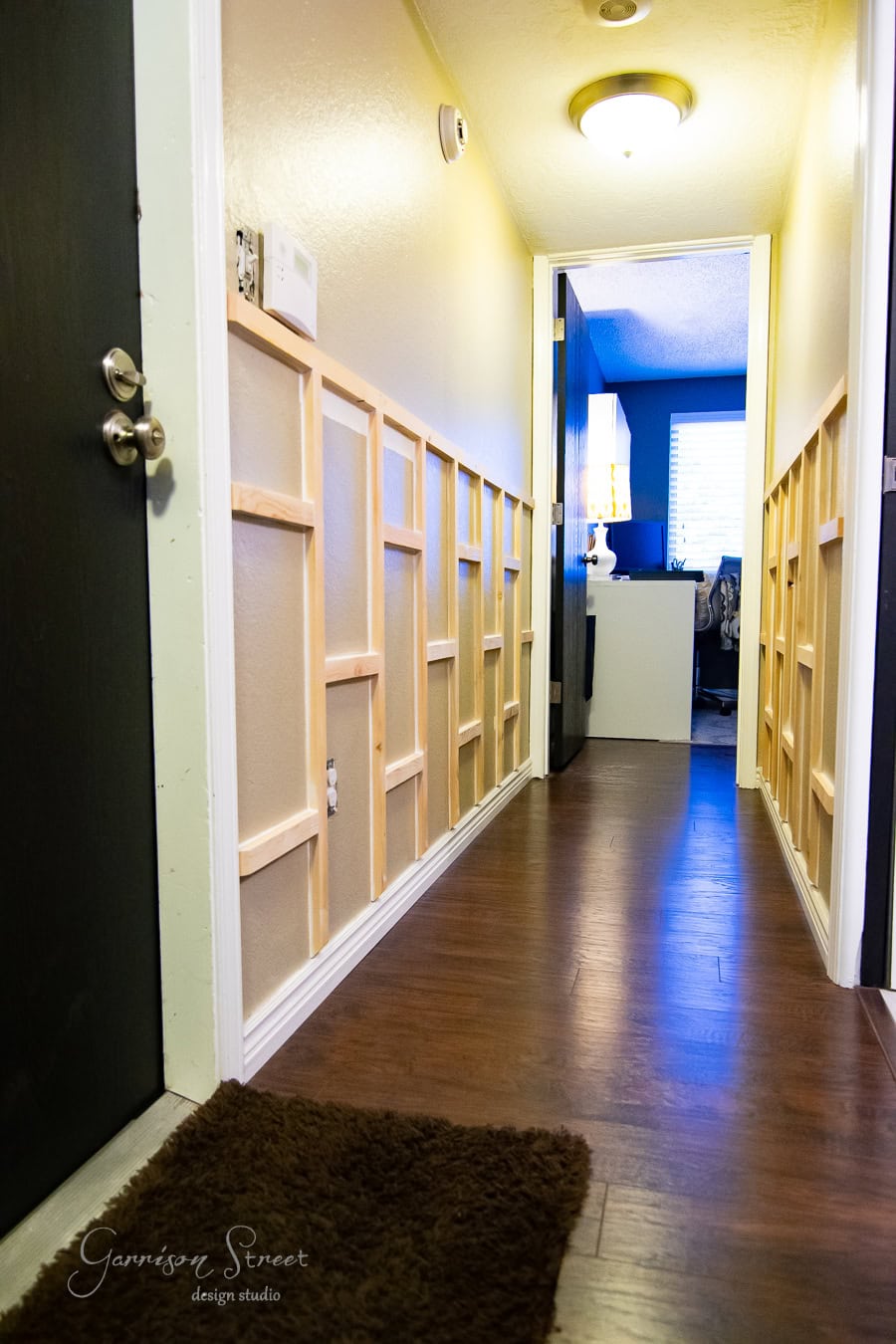 A hallway with board and batten paneling on the walls and dark wood flooring. A door is open on the left, revealing a warm glow from a lit room at the end of this inviting DIY-enhanced hall.
