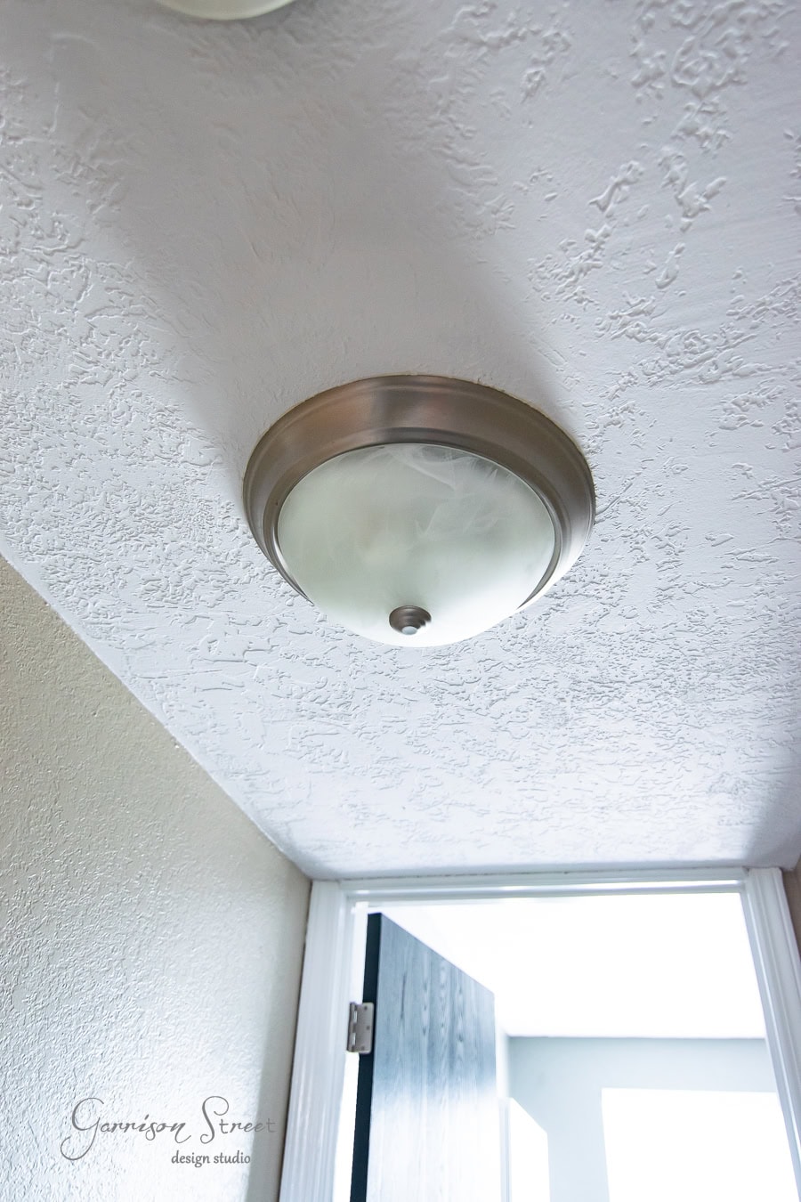 A close-up view of a ceiling light fixture with a frosted glass cover, mounted on a textured white ceiling in the hall. Below, part of an open black door is visible, with sunlight streaming in from a window or door opening, enhancing the board and batten detail on the walls.