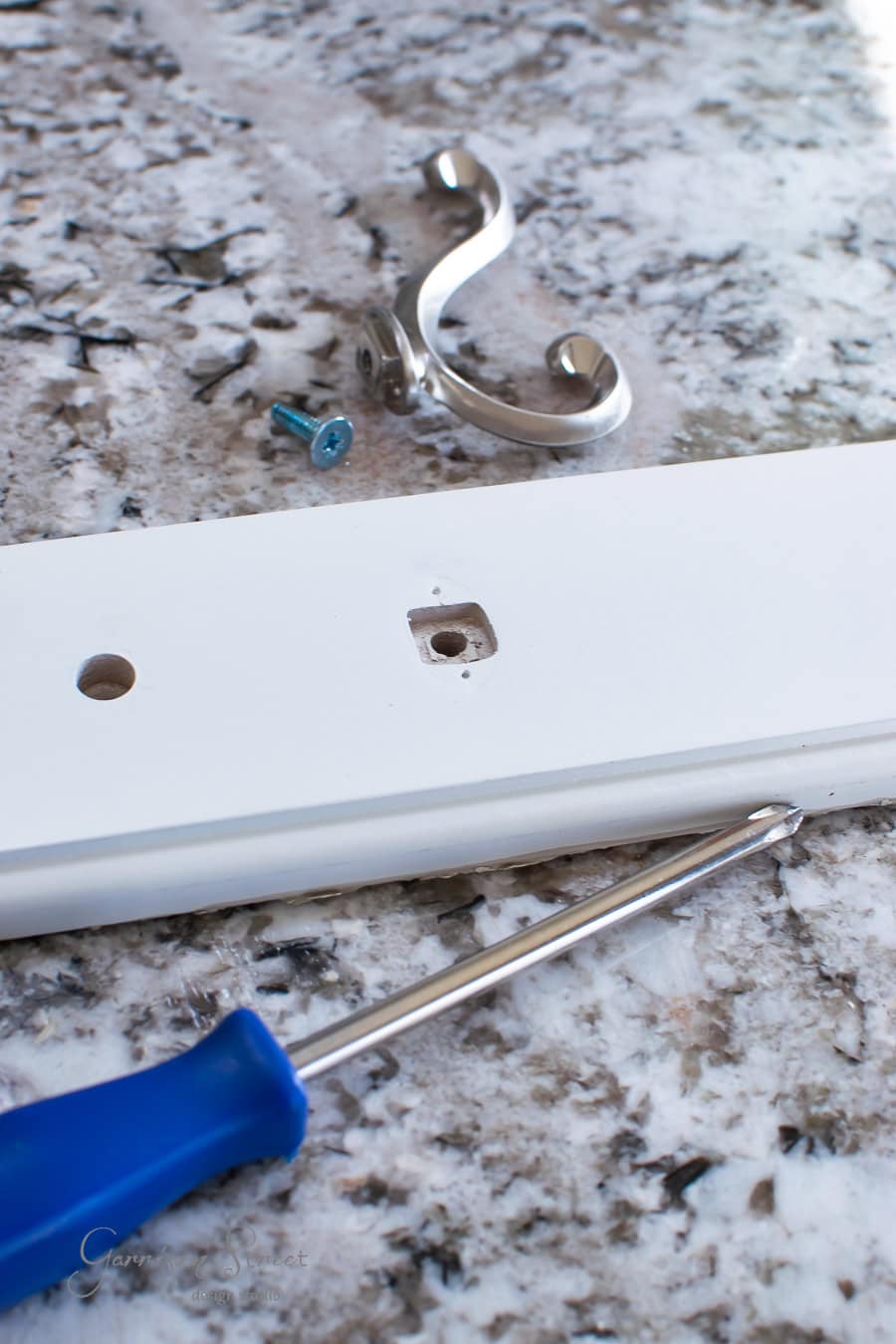 On a speckled granite countertop, a blue-handled screwdriver rests beside a white wooden board with a drilled hole, ideal for your DIY hall project. Nearby, a silver metal hook and two screws await assembly for that perfect board and batten look.