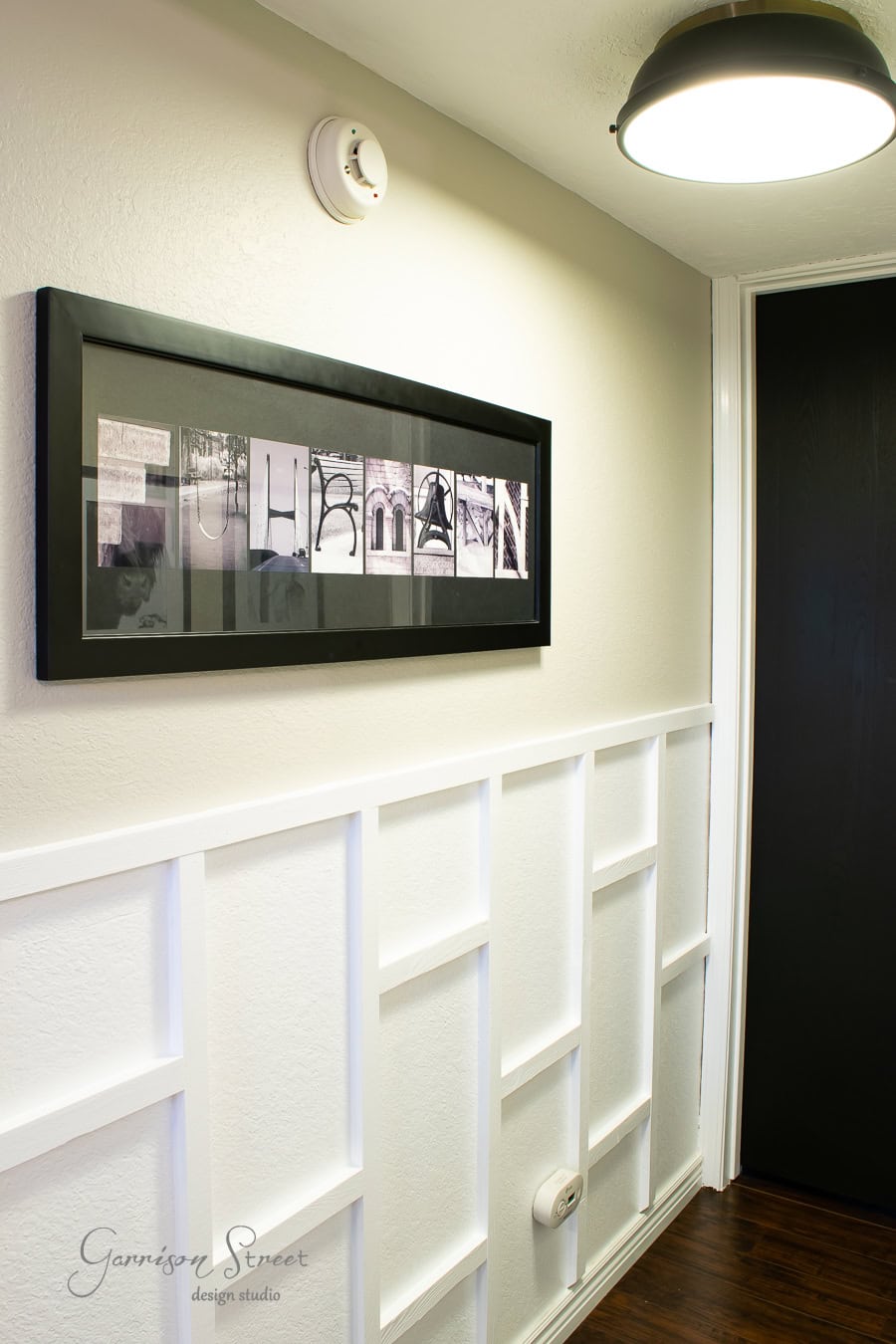 The hall boasts a DIY touch with white panelled board and batten walls complemented by dark wood flooring. A rectangular framed art piece with abstract designs adorns the space, while a ceiling light and smoke detector are subtly visible above.
