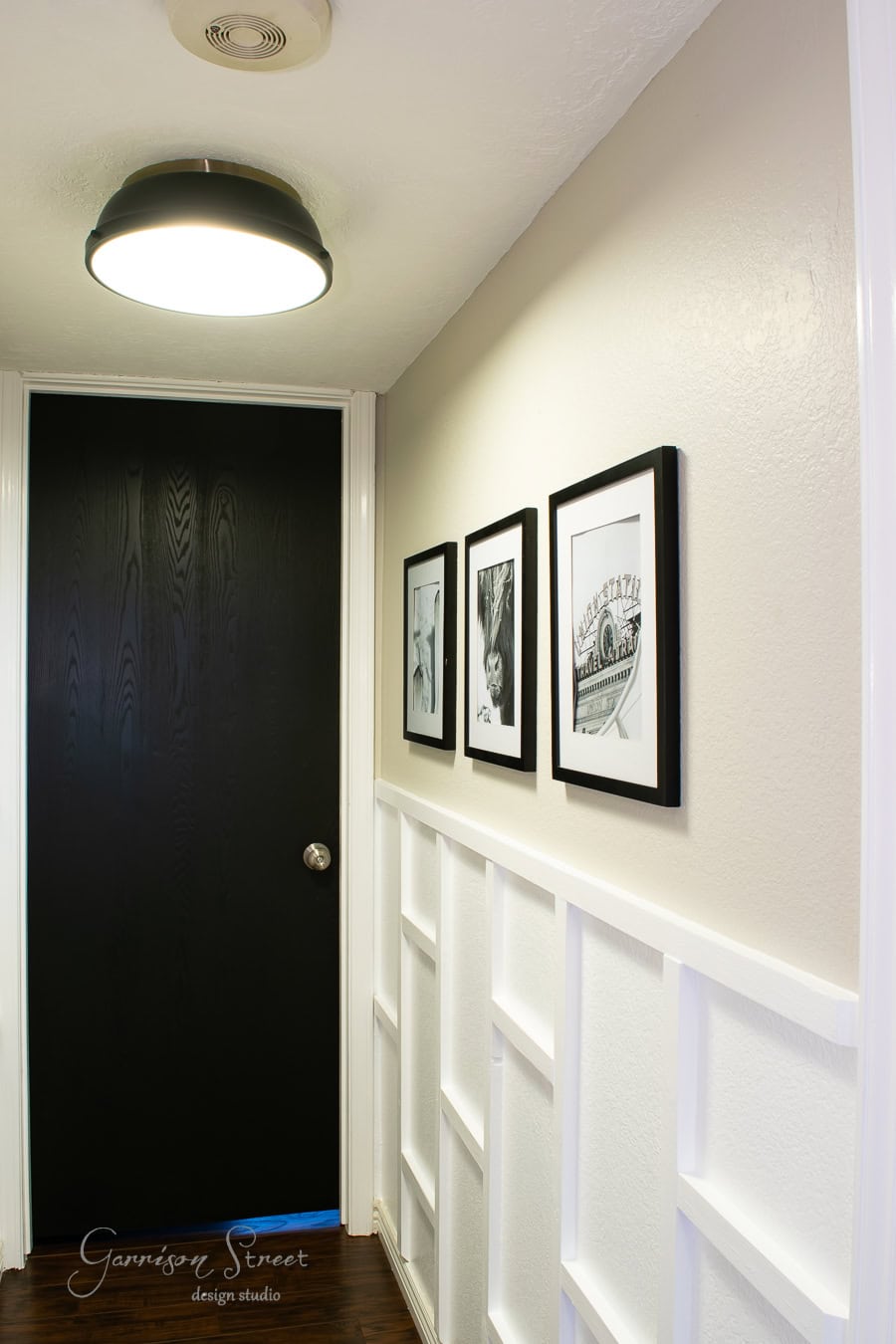 A narrow hallway with a dark door at the end, illuminated by a ceiling light. The walls are painted light beige, adorned with three framed black and white photos on the right. DIY board and batten adds texture to the lower half of the walls, enhancing its classic charm.