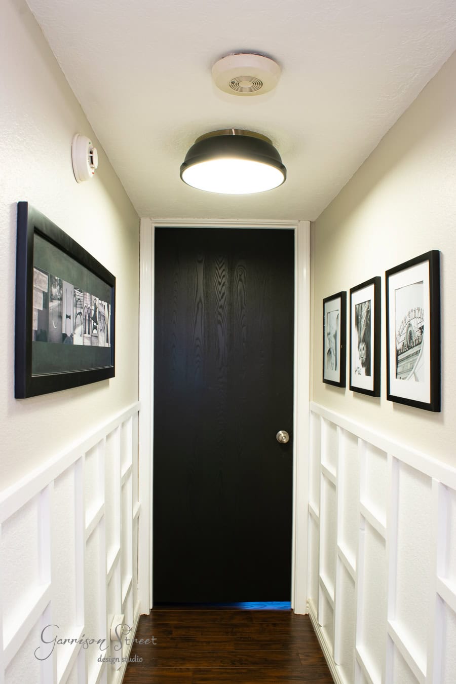 The narrow hallway, with its board and batten walls, features black and white framed photos elegantly displayed on both sides. A round ceiling light illuminates the space leading to a dark wooden door with a silver doorknob, while dark wooden floorboards complete this DIY-inspired hall.