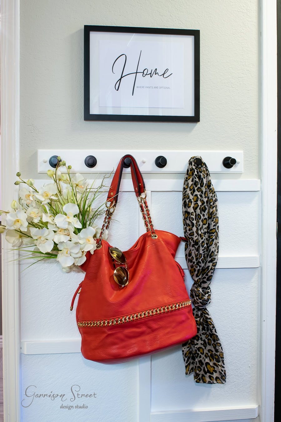 A wall-mounted coat rack against a DIY board and batten wall features a framed "Home" sign above. A vibrant red handbag with chain straps, sunglasses, a floral bouquet, and a leopard print scarf hang on the hooks. The logo for Garrison Street Design Studio adorns the bottom.