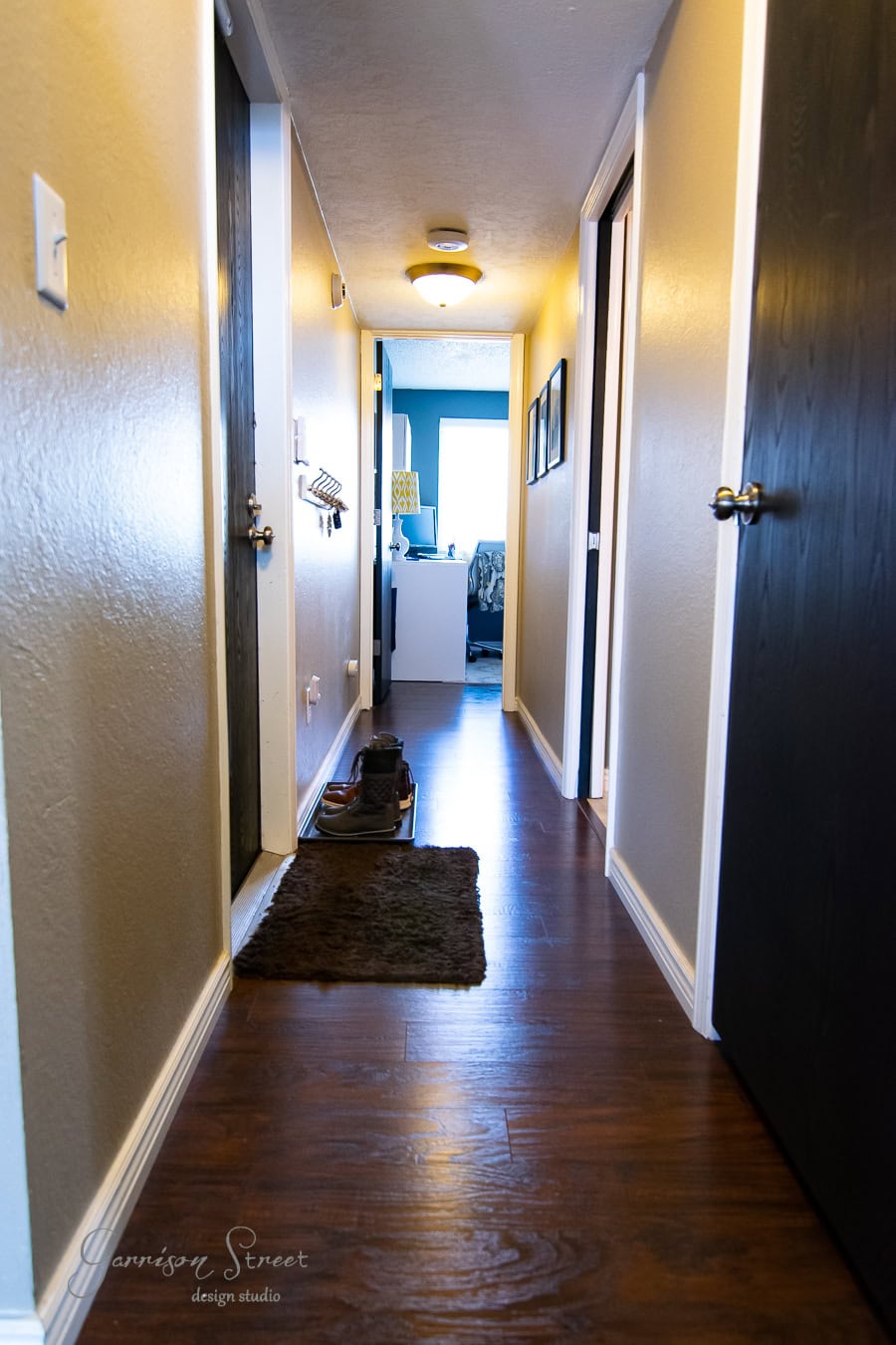 A narrow hall with dark wooden floors leads to a bright room. Shoes rest on a mat against one wall. The beige walls feature DIY board and batten designs, with closed doors on either side and a ceiling light fixture. A glimpse of an office setup is visible at the end.