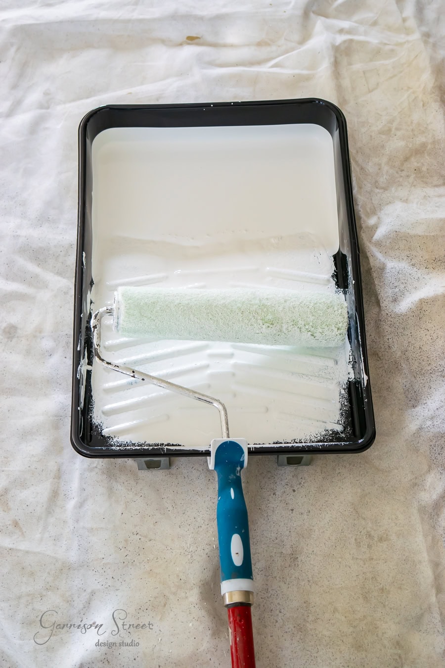 A paint roller with a blue handle rests in a black tray filled with white paint, set on a white, textured sheet. This setup hints at a makeover project unfolding, possibly during quarantine, transforming spaces one stroke at a time.