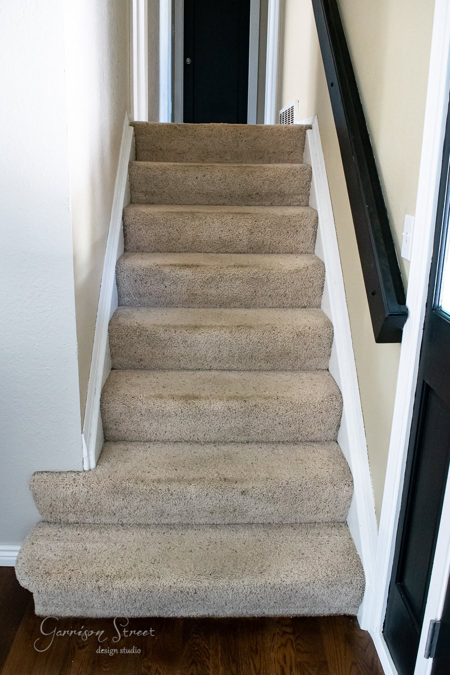 A staircase with beige carpet and a black handrail on the right leads to a narrow, light-colored hallway with a dark door at the top. This project, crafted by Garrison Street Design Studio, features a subtle makeover that transforms simplicity into elegance.