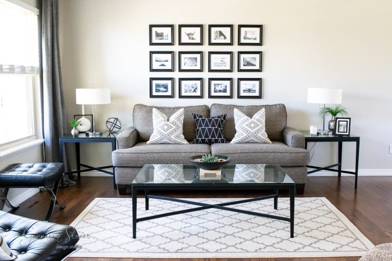A modern living room with a gray sofa, patterned pillows, and a glass coffee table on a patterned rug. The wall features a grid of black-and-white framed photos. Two black side tables hold lamps and small plants. Natural light brightens the space.