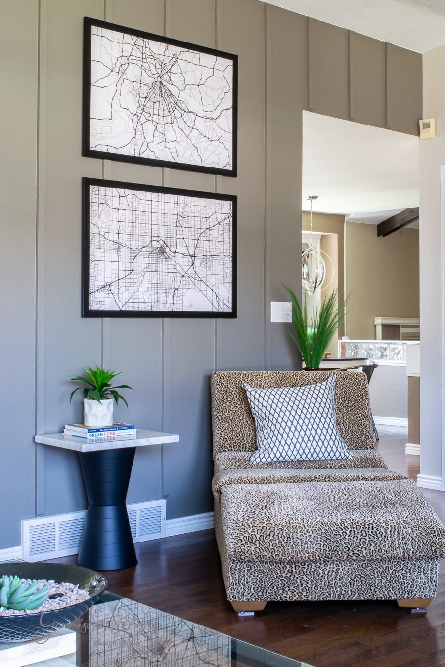 Cozy living room with a patterned chaise lounge, a geometric pillow, and decorative plants on a round side table. Two framed map prints hang on a gray paneled wall. A quarantine makeover project brought sunlight to brighten the space, highlighting the wooden floor.