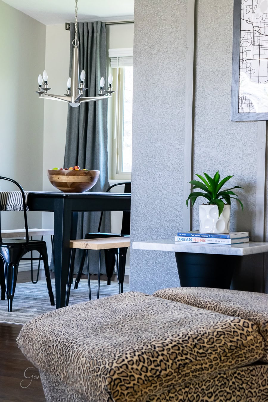 Modern dining area with a wooden table, black chairs, and a bowl of fruit. A chandelier hangs above, and a plant sits on a side table with books. As part of the recent makeover project, leopard-print seating is in the foreground with a window and gray curtains in the background.