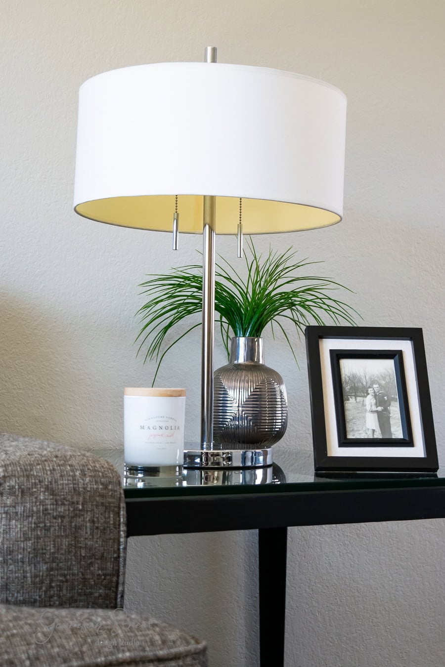 A modern table lamp with a white shade sits on a black side table—a perfect touch in this quarantine makeover. Nearby, a silver vase holds green foliage beside a candle and framed black-and-white photo. A corner of a gray sofa is visible against the beige wall, completing the project.