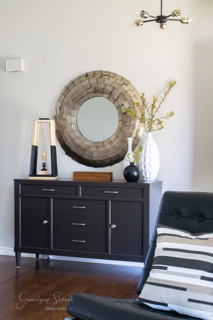 In a recent makeover project, a stylish room features a dark wood sideboard with a large decorative round mirror above it. On the sideboard are a modern lamp and a tall white vase with branches. A black leather chair with a patterned pillow sits to the side, beneath a contemporary light fixture.