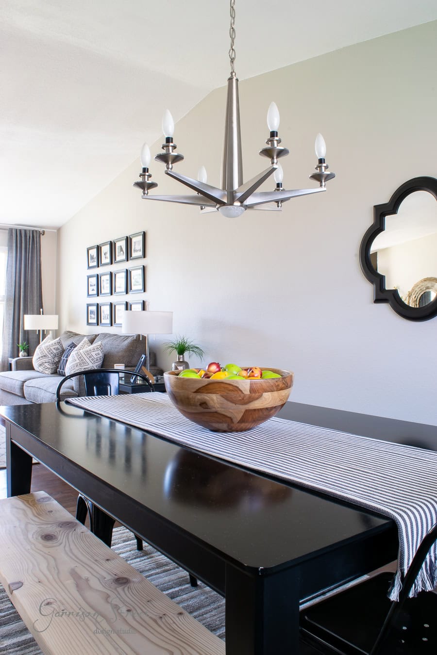 A dining room makeover showcases a black table with a striped runner, topped with a wooden bowl of colorful fruit. Overhead, a modern chandelier adds elegance. The background reveals a mirror, sofa, and framed pictures—a perfect quarantine project turned into an inviting retreat.