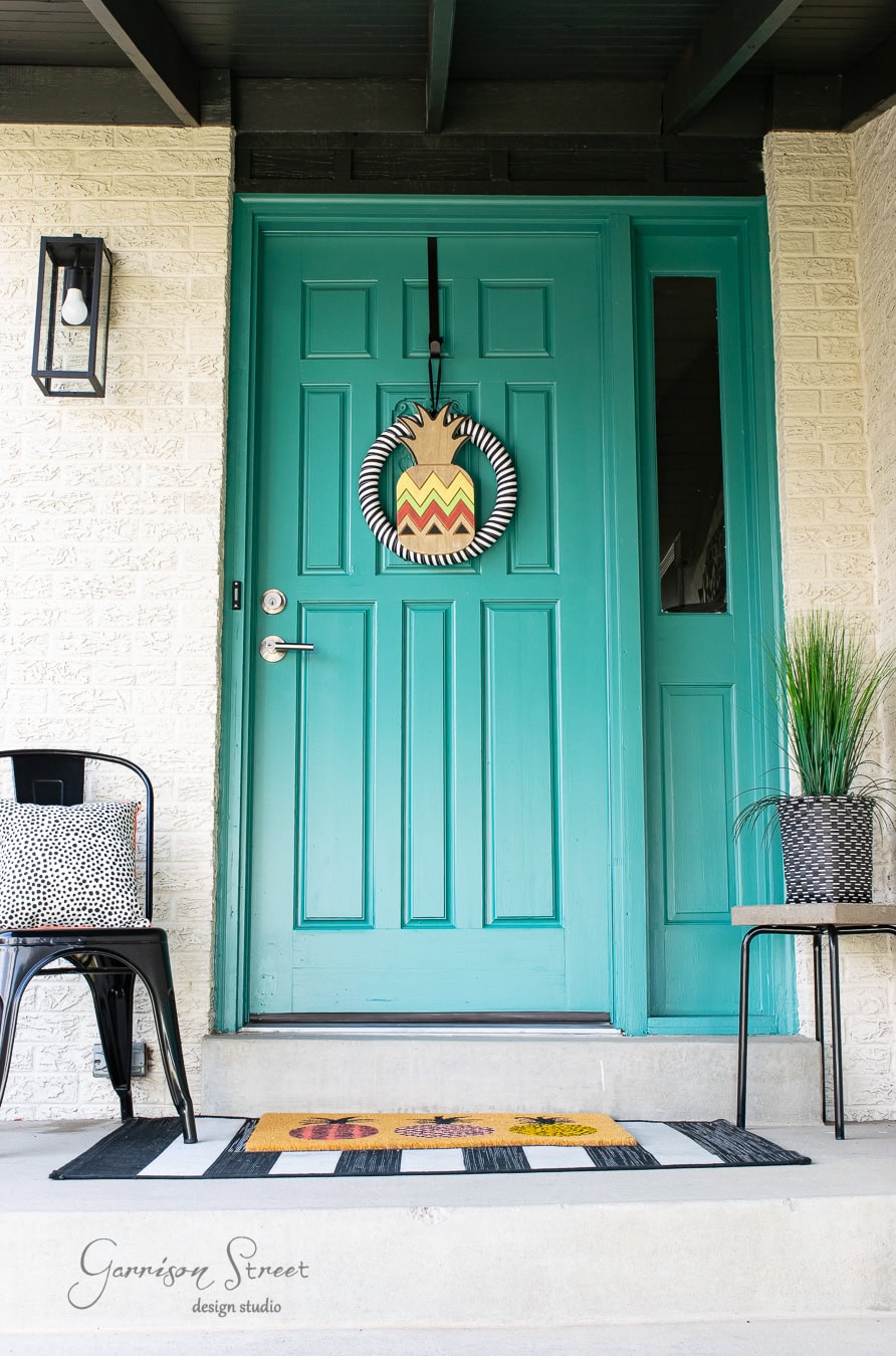 A teal front door adorned with a vibrant wreath, showcasing spring charm with black and white stripes and zigzag patterns. The front porch features a black and white striped mat with floral decor, accompanied by a cozy chair with a pillow and a pot of tall grass.