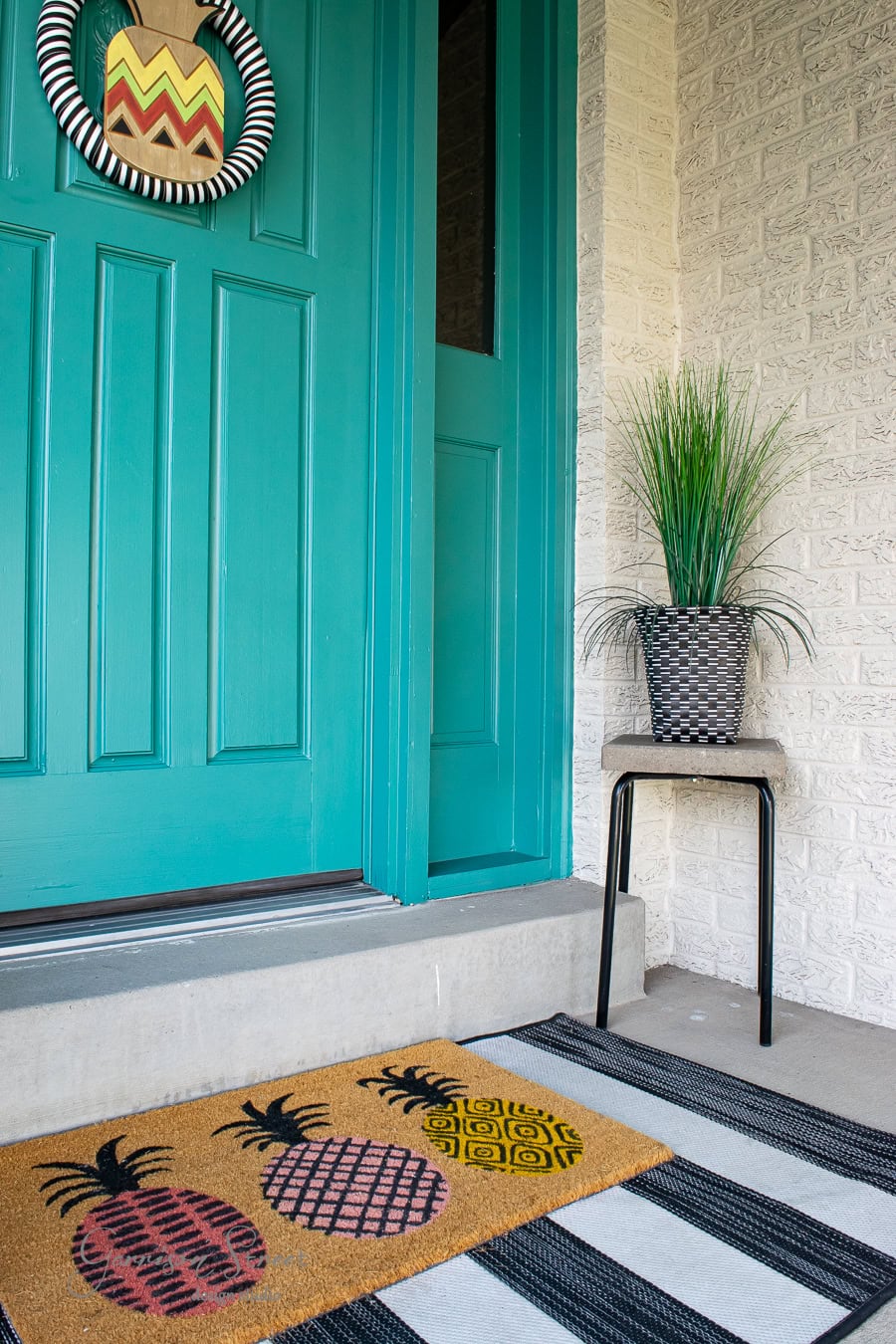 A teal front door with a decorative wreath featuring a pineapple design welcomes you to this charming spring decor. On the striped rug, a coir doormat adorned with colorful pineapples adds cheer, while a small black table holds a potted plant on the inviting front porch.