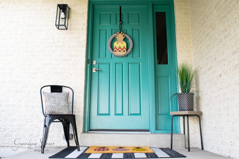 A teal front door with a decorative wreath opens onto a porch. A black chair with a patterned cushion sits beside the door, and a potted plant stands on the other side. A colorful doormat lies on the ground.
