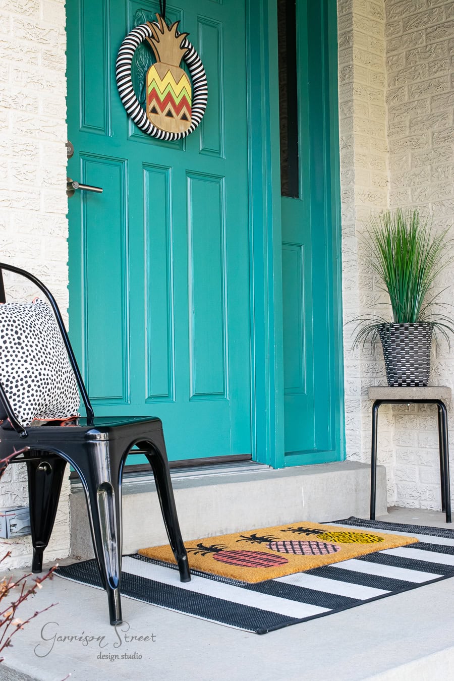 A vibrant turquoise front door with a pineapple wreath welcomes spring. A black metal chair with a polka-dot cushion sits on the decor-rich front porch, boasting a black-and-white striped rug. A potted plant on a small table adds a touch of greenery.