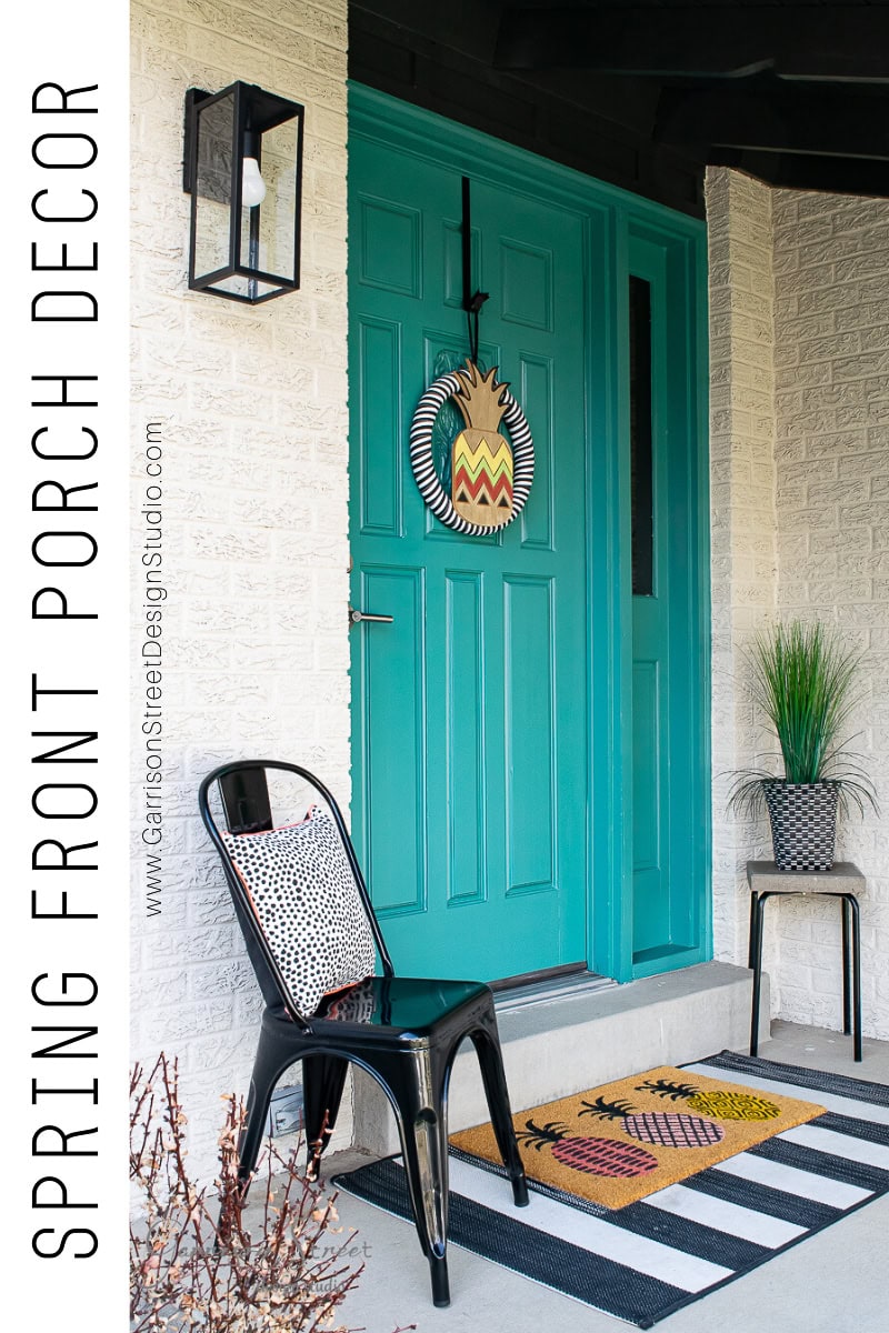A teal front door adorned with a decorative wreath sets the tone for spring decor, featuring two black chairs with patterned cushions and a potted plant. A striped doormat welcomes visitors on the front porch. A modern wall light illuminates the text "Spring Front Porch Decor" on the side.