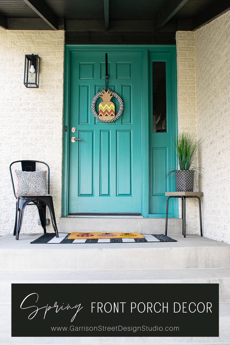 A teal front door adorned with a chevron wreath and modern house number on cream brick. A black chair with a patterned cushion and a plant on a small stand flank the entryway. The vibrant scene is completed by "Spring Front Porch Decor" text and a colorful mat, embodying delightful spring vibes.
