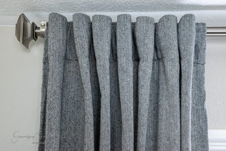 Close-up of gray herringbone-patterned curtains hanging on a silver curtain rod with a decorative square finial. The fabric has a structured, pleated design.