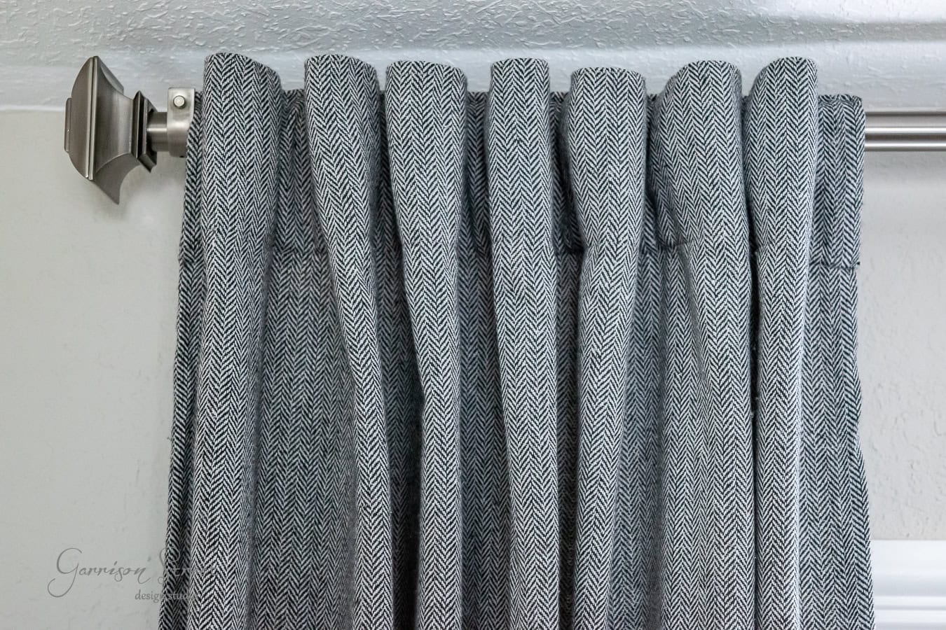 Close-up of gray herringbone-patterned curtains hanging on a silver curtain rod with a decorative square finial. The fabric has a structured, pleated design.
