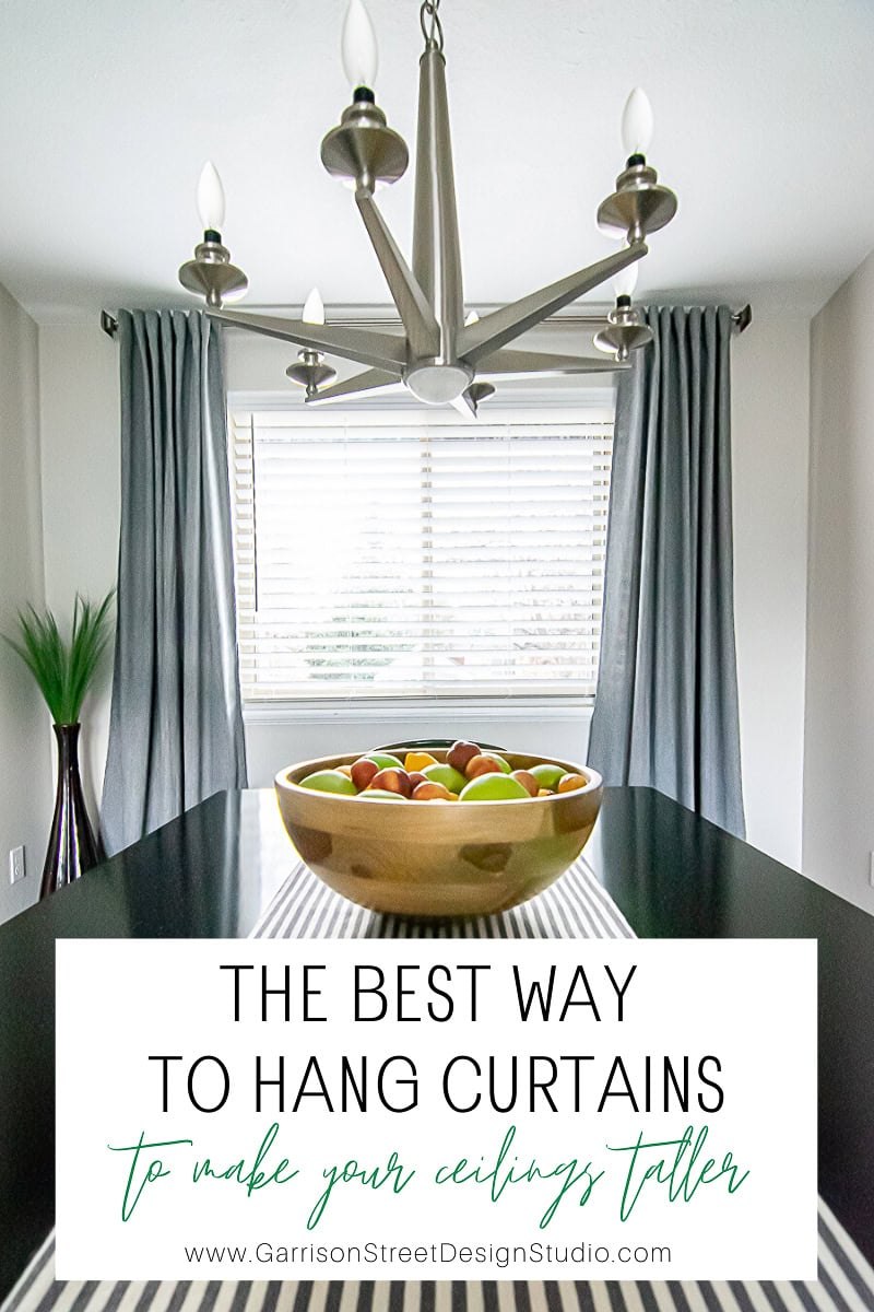 A modern dining room showcases a wooden bowl filled with fruit on the table. A silver chandelier complements the long curtains hanging elegantly beside the window. Text overlay reads, "Discover the best way to hang curtains and make your ceilings appear taller.