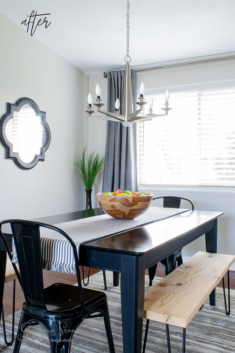 A modern dining room features a black table, wooden bench, and sleek black chairs. A wooden bowl with fruit elegantly sits on the table. The silver chandelier hangs as the best focal point above a rectangular rug, while curtains frame the mirror and plant on white and gray walls.